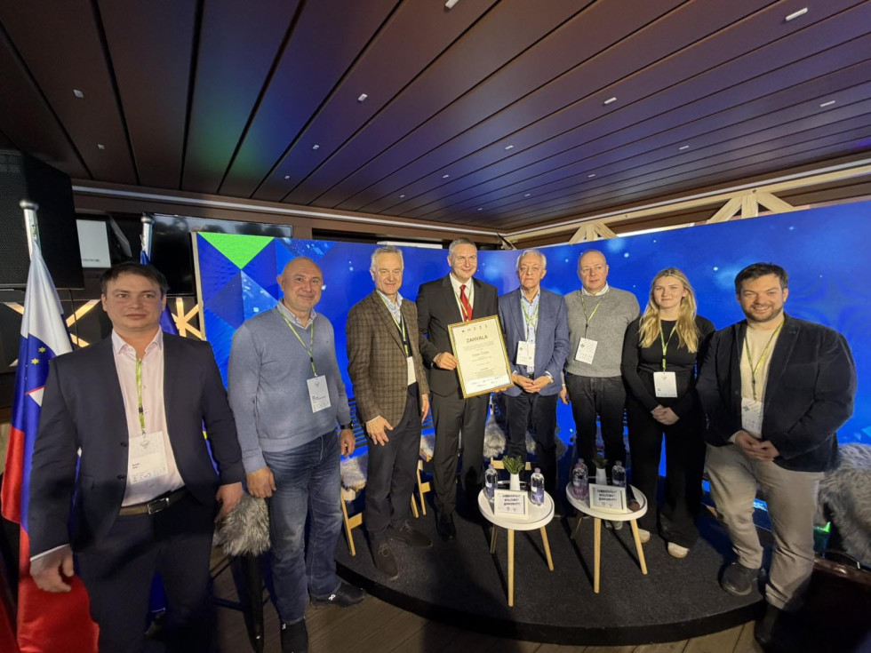 Sedem moških in ženska stojijo eden zraven drugega. V ozadju je velik svetlomoder LED-zaslon. Ob njem slovenska zastava. 