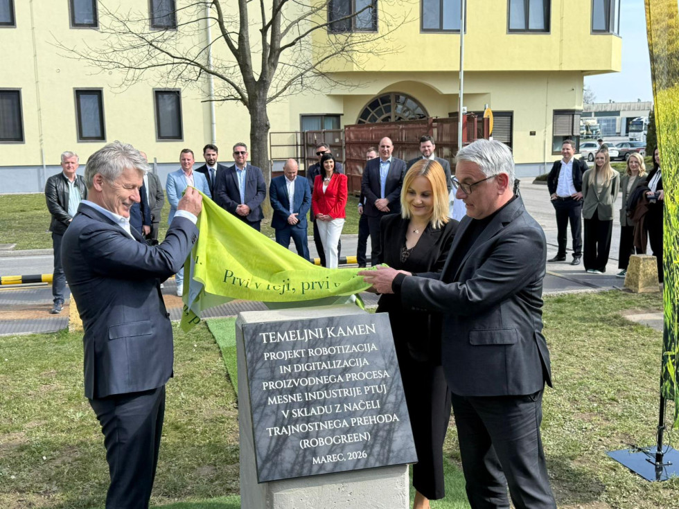 Dva moška odkrivata temeljni kamen. Nedaleč stran stojijo opazovalci. 