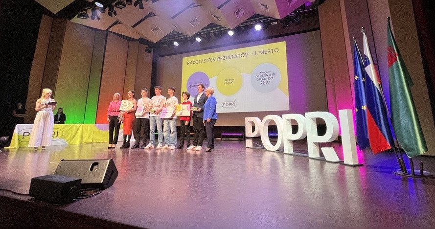 Na odru stojijo mladi, predsednica države in moški. V ozadju na velikem platnu predstavitev. Na odru velike črke, ki tvorijo napis Popri, zraven črk zastave. 