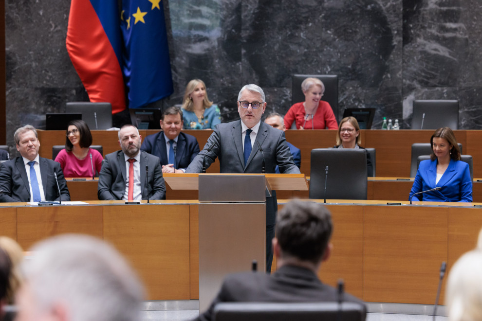 minister Han at speaking table in National Assembly of Republic of Slovenia with other ministers seating behind him.