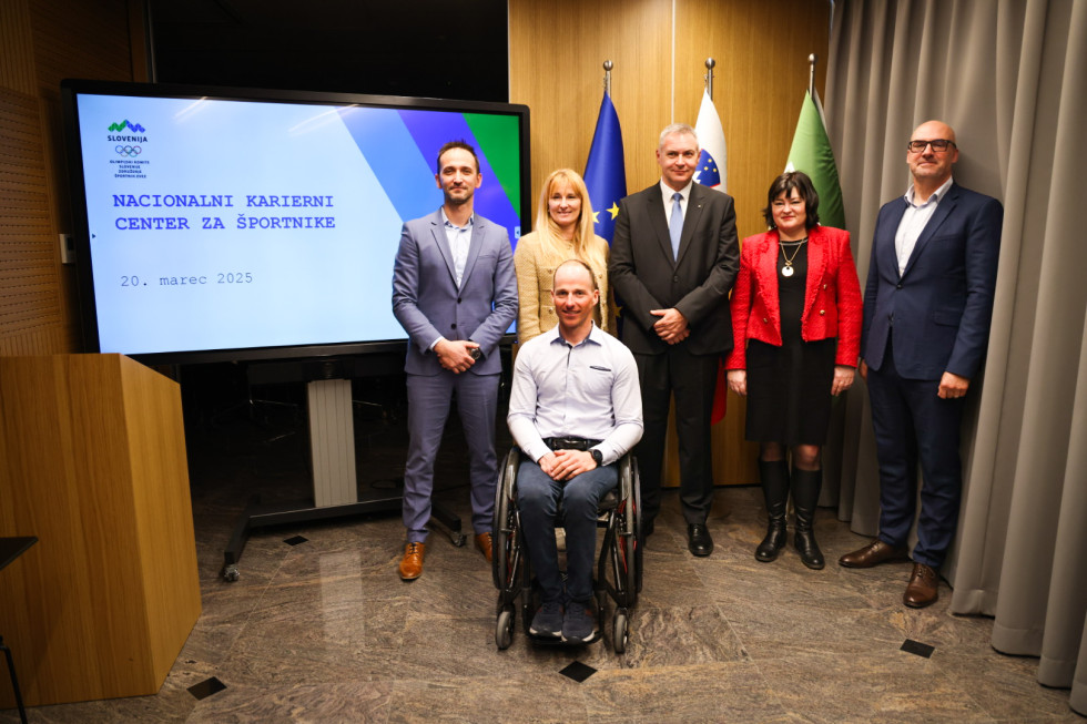 Skupna slika. Moški in ženske stojijo, spredaj gospod na vozičku. Zadaj zastave in velik ekran z napisom: Nacionalni karierni center za športnike.