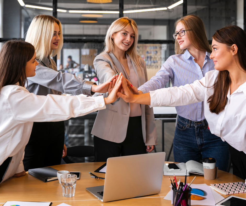 pet žensk stoji za delovno mizo s sklenjenimi dlanmi v znak skupinskega dela ali podpore