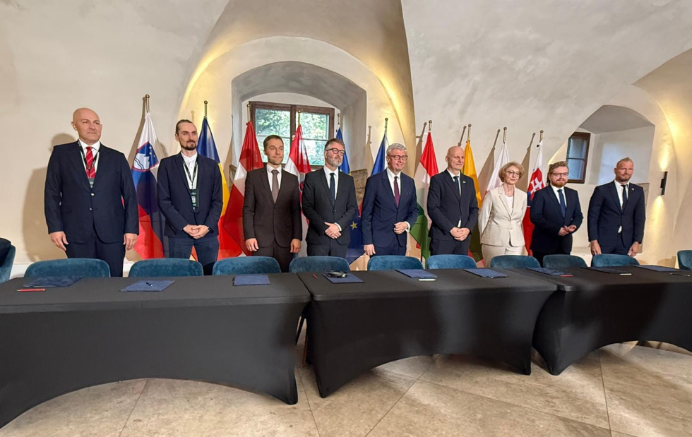 Ministri več držav stojijo za mizo s podpisnimi mapami pred državnimi in evropsko zastavo tik pred podpisom Praške deklaracije o umetni inteligenci.