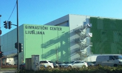 Fotografija stavbe Gimnastičnega centra Ljubljana od zunaj. Pred gimnastičnim centrom so avtomobili na mimoidoči cesti.