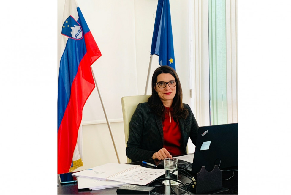 Ministrica za računalnikom v sejni sobi ministrstva. V ozadju slovenska in evropska zastava.