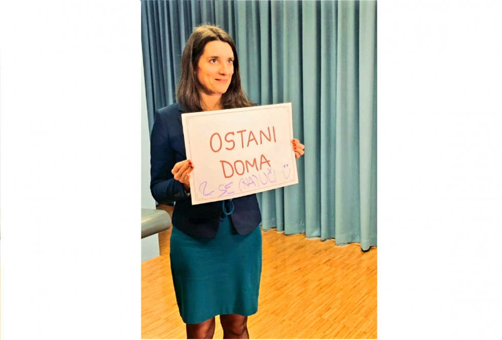 Ministrica v tiskovnem središču drži tablo z napisom 'Ostani doma'.