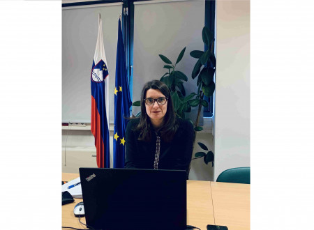  Ministrica za računalnikom in gleda v objektiv. V ozadju zastava Slovenije in EU.