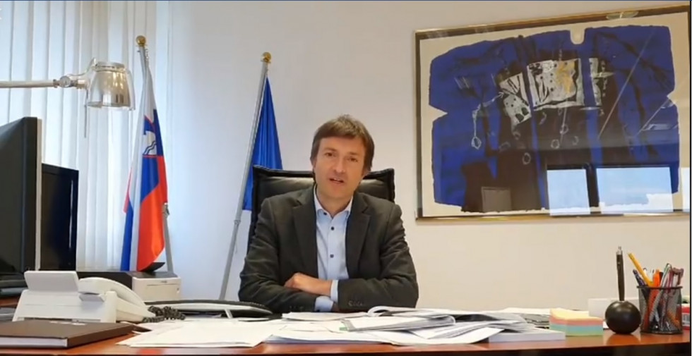 Videonagovor državnega sekretarja, ki sedi za pisalno mizo v svoji pisarni. V ozadju slovenska in evropska zastava.