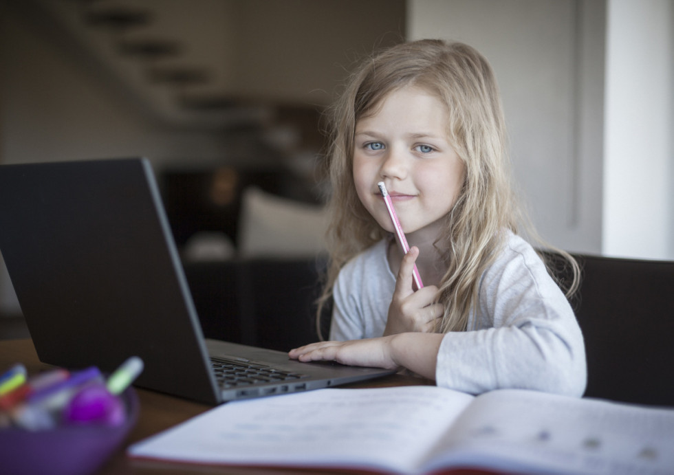 Učenka osnovne šole se doma za mizo z laptopom uči na daljavo.