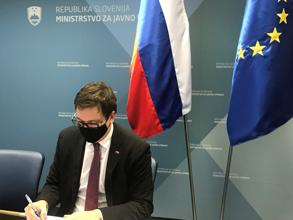 Minister Koritnik sedeč za mizo, pred njim računalnik, v ozadju zastavi