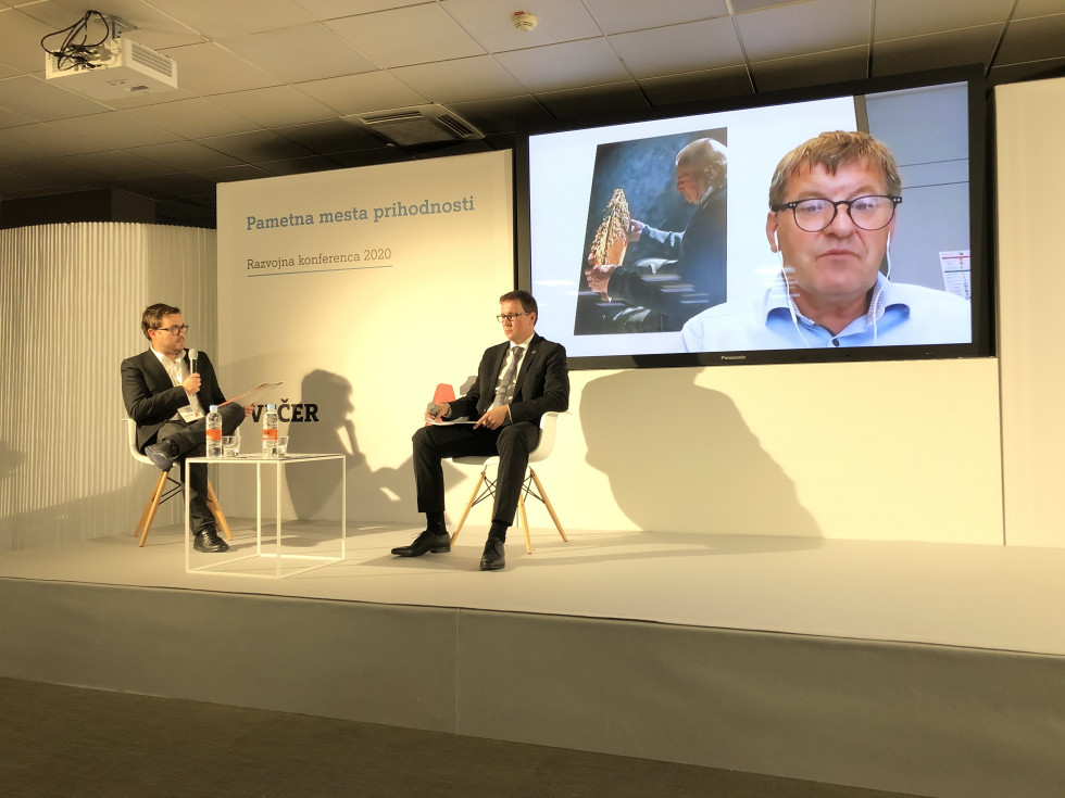 moderator in minister sedita na odru, v ozadju na ekranu slika evropskega poslanca Bogoviča, ki sodeluje prek video povezave