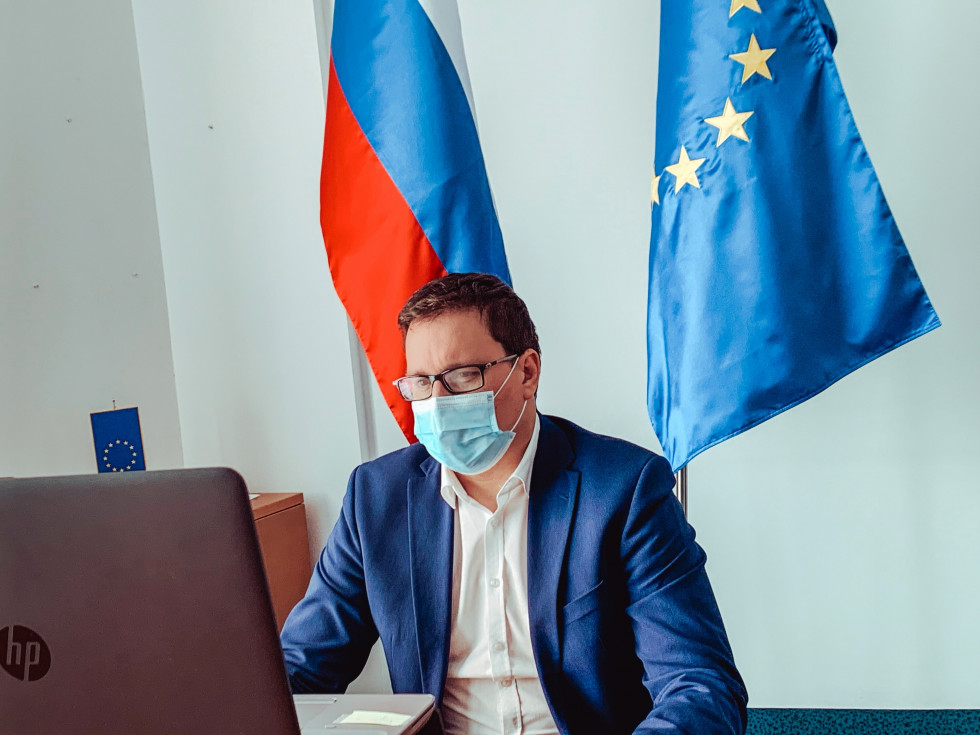 the minister is sitting in front of the screen, in the background the Slovenian flag and the flag of the European Union