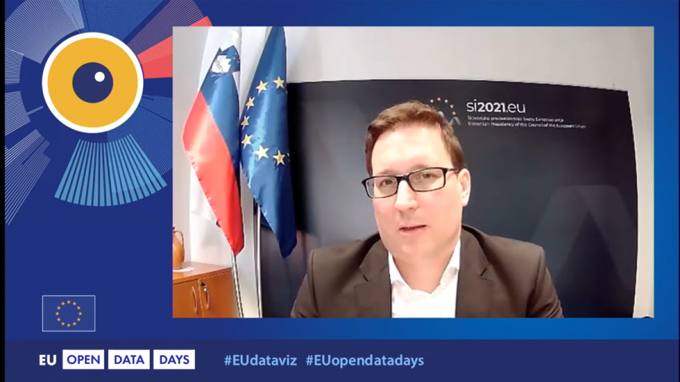 Zaslon, na kateri ima nagovor minister, zato je vidna njegov obraz, v ozadju pa slovenska in EU zastava in modri pano z logotipom Ministrstva za javno upravo