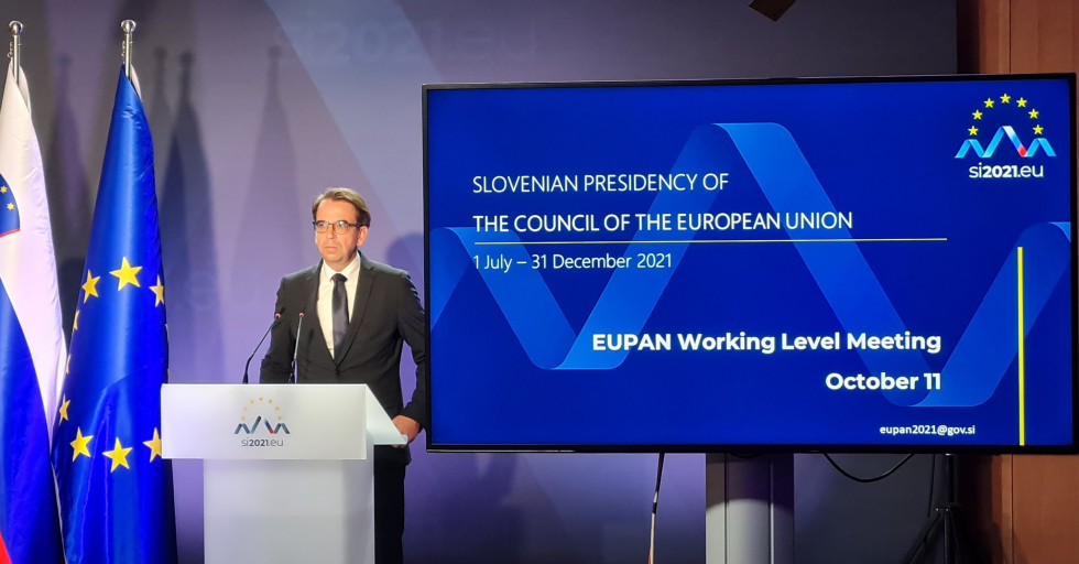 Direktor direktorata stoji pred govorniškim pultom, zadaj ekran na katerem je naslov konference, levo od govorca slovenska in EU zastava.