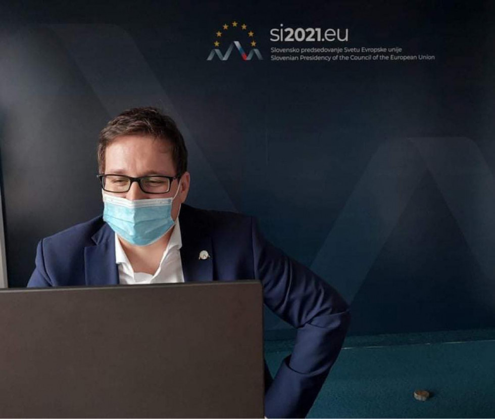 Minister Koritnik sedeč za računalnikom, za njim ozadje predsedovanja