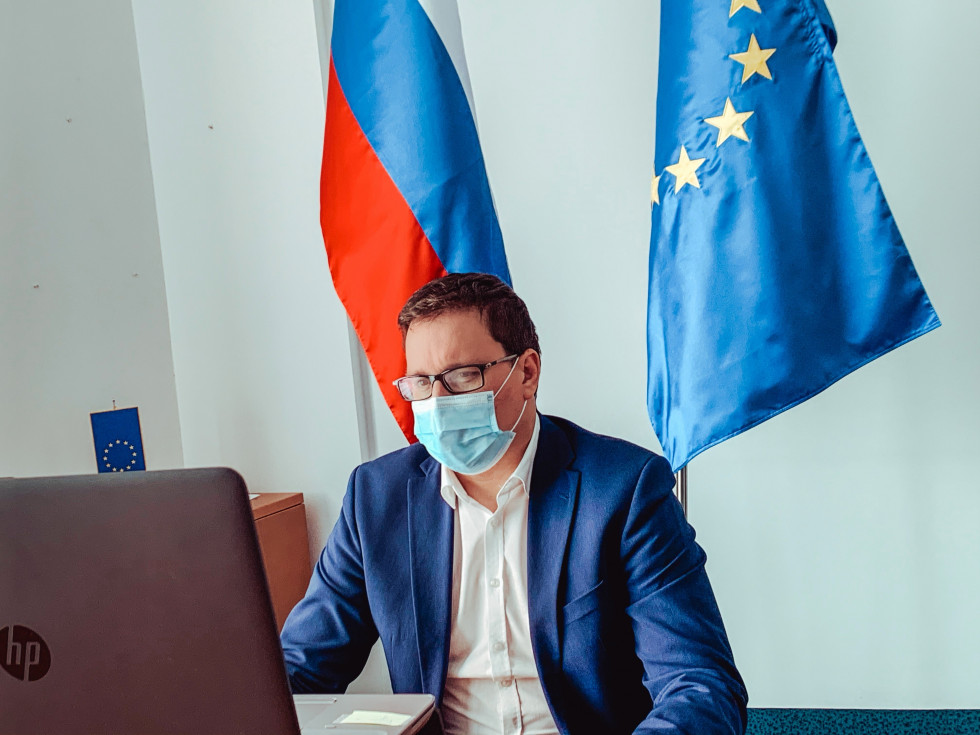 minister sedi pred ekranom z zaščitno masko na obrazu, v ozadju slovenska zastava in zastava Evropske unije