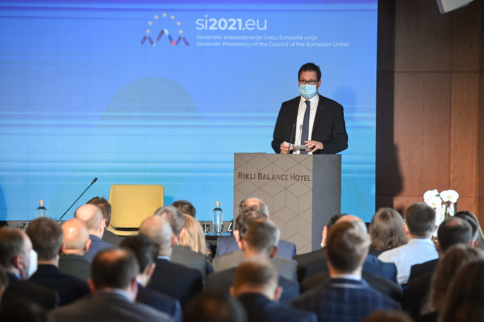 Minister Koritnik stoji za govornico, v ozadju je na platno projecirano modro ozadje z logotipom predsedovanja Slovenije