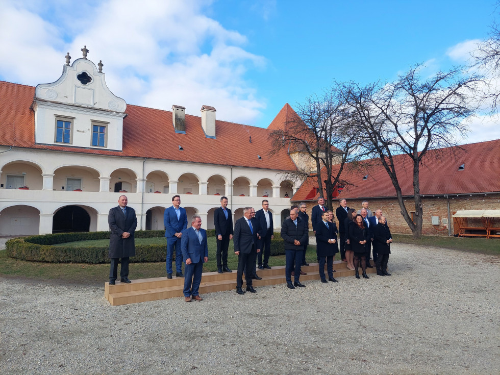 Skupinska fotografija članov vlade. Člani stojijo na dveh lesenih stopnicah, v ozadju dvorec Rakičan