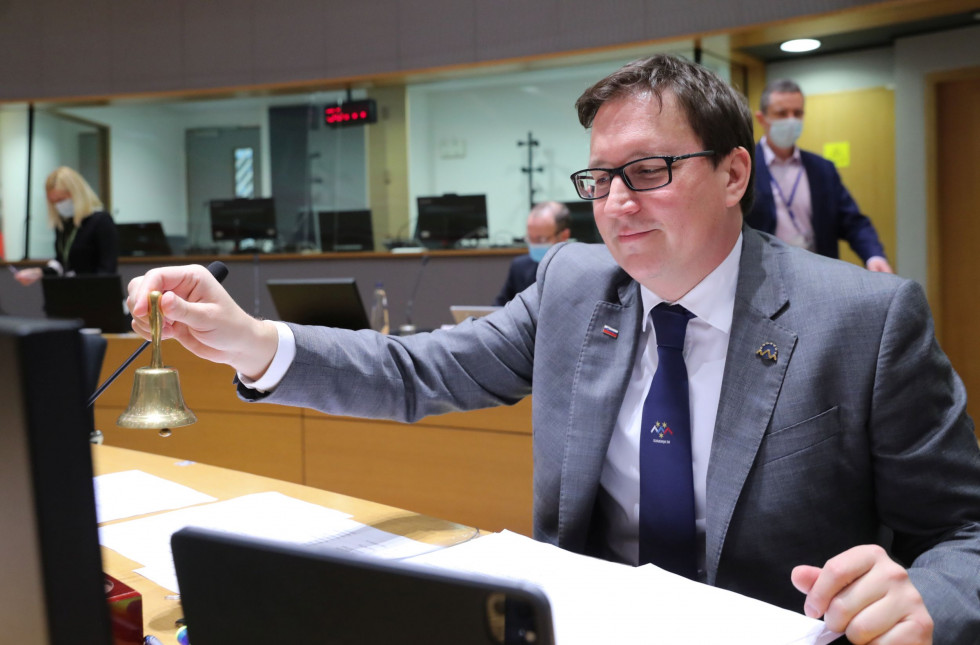 Minister sedi za mizo v kongresni sobi, pred njim računalnik, v roki drži zvonček za začetek zasedanja.