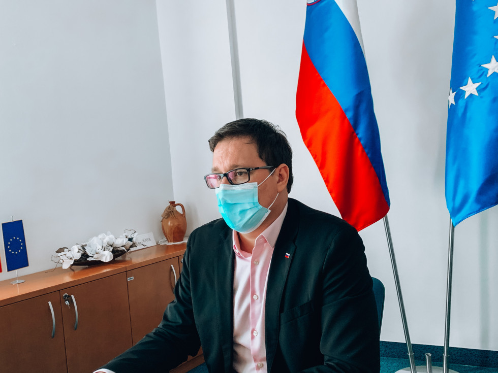 minister sedi za računalniškim zaslonom , v ozadju slovenska in evropska zastava