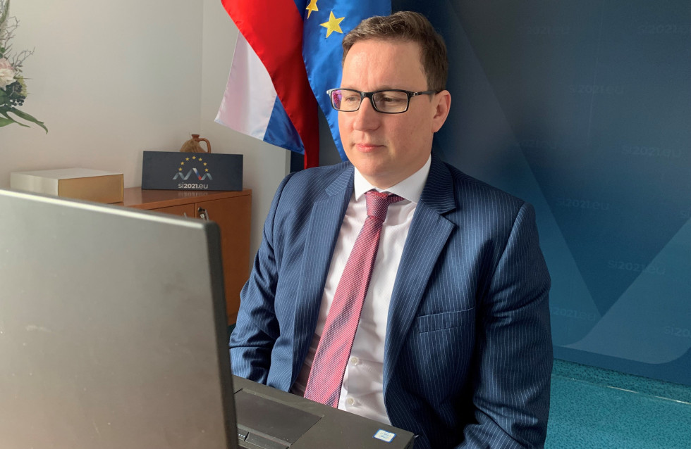 fotografija ministra, ki sedi v sejni sobi pred računalnikom. V ozadju slovenska in UE zastava in pano ministrstva.