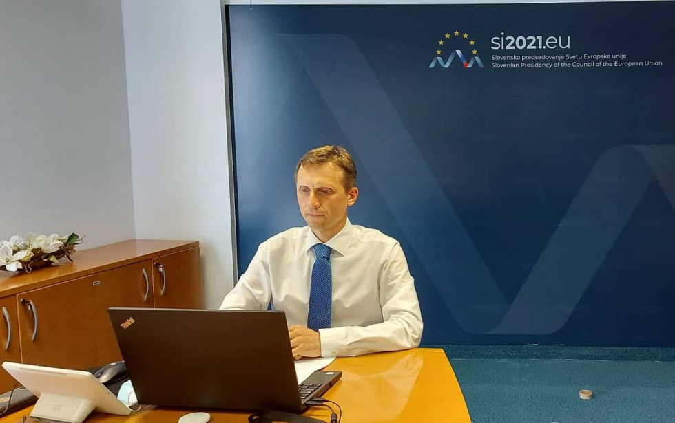 državni sekretar sedi za mizo pred računalniškim monitorjem, v ozadju modri pano Ministrstva za javno upravo in predsedovanja EU