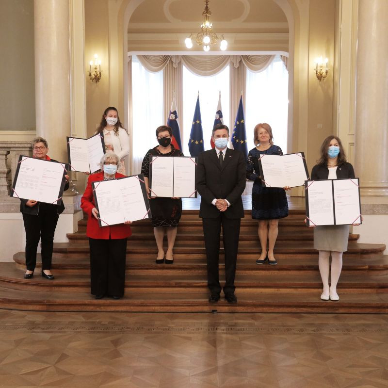 prejemniki priznanj stojijo na stopnicah v veliki dvorani predsedniške palače, v rokah držijo odprte mape s priznanji, v sredini je predsednik republike, v ozadju pa zastave Slovenije in Evropske unije