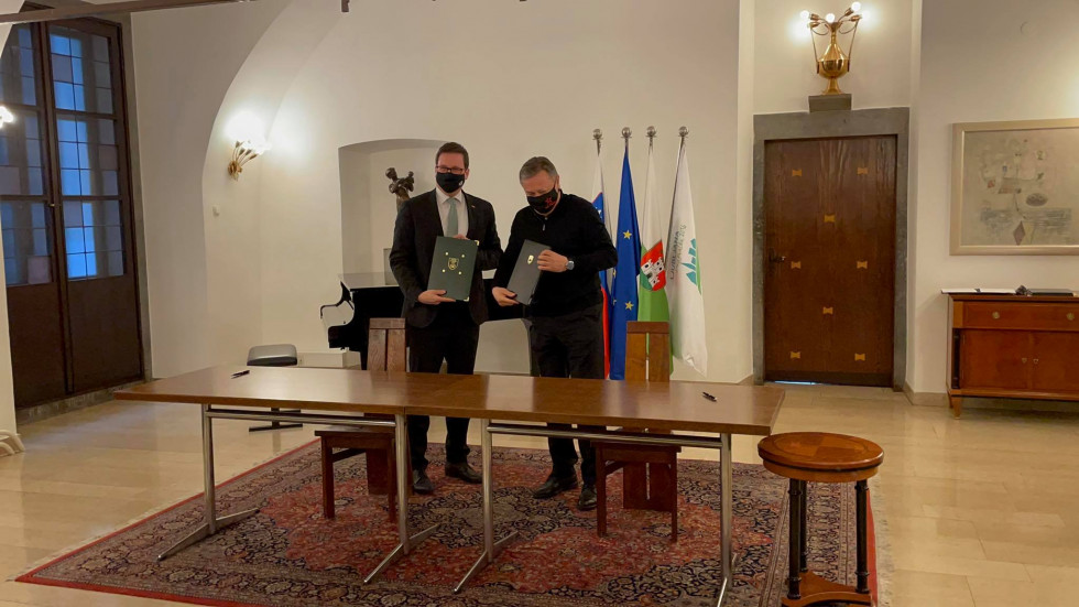 stoječa fotografija, podpisnika dogovora, minister in župan, stojita za mizo z dvignjenimi mapami, črne barve, v katerih je dogovor (imata zaščitni maski)