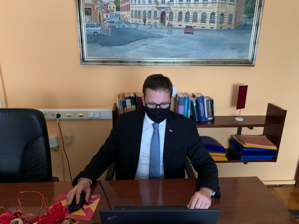 Minister pred zaslonom računalnika sede spremlja zasedanje in se ga aktivno udeležuje. V ozadju knjižna polica s knjigami in slika