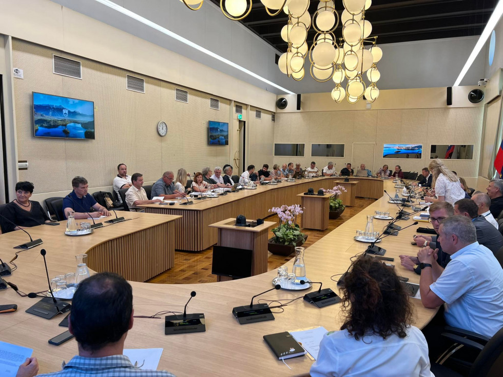 Udeleženci seje sedijo za ovalno veliko mizo (cca 80 oseb), pred njimi so namizni mikrofoni, v ozadju s stropa visi lestenec, vidni so ekrani na stenah v dvorani