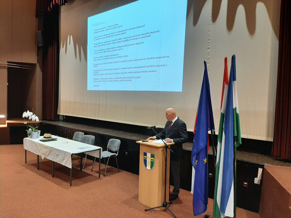Govorec stoji pred govorniškim pultom, v ozadju prezentacija in slovenska, EU zastava in zastava občine gostiteljice
