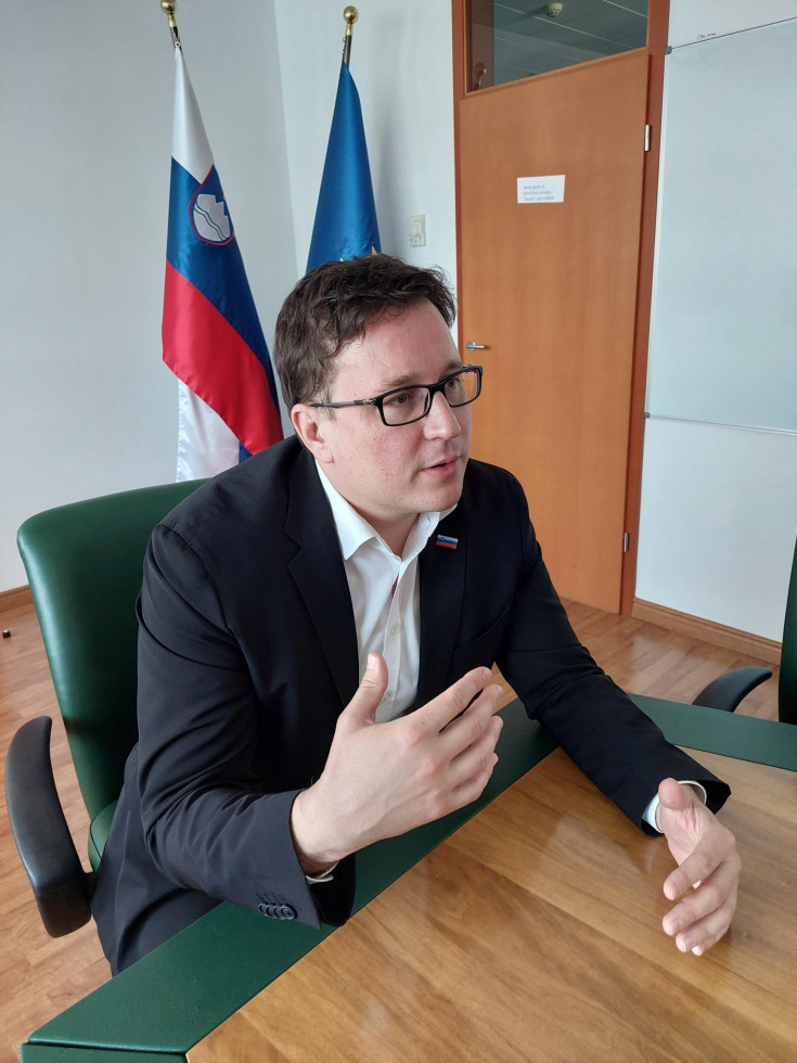 Minister sedi na stolu pred ekranom in nagovarja na video dogodku. Za njim obe zastavi slovenska in EU.