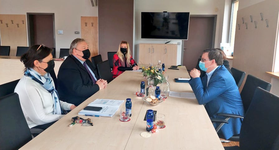 Minister in župan s sodelavci sedijo za svetlo rjavo mizo, na steni LCD monitor, na mizi cvetlični aranžma, voda in sok. Sogovorniki imajo zaščitne maske.