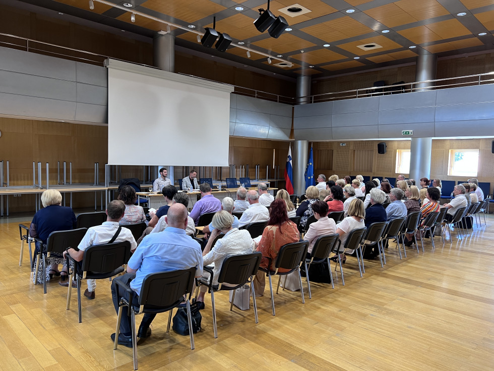 V dvorani s hrbtne strani slikani udeleženci srečanja, vidni so stoli, ljudje obrnjeni stran od kamere, vsi gledajo v ospredje dvorane, kjer stoji miza, za katero sedita predstavnika Ministrstva za javno upravo, v ozadju je vidno platno za projekcijo