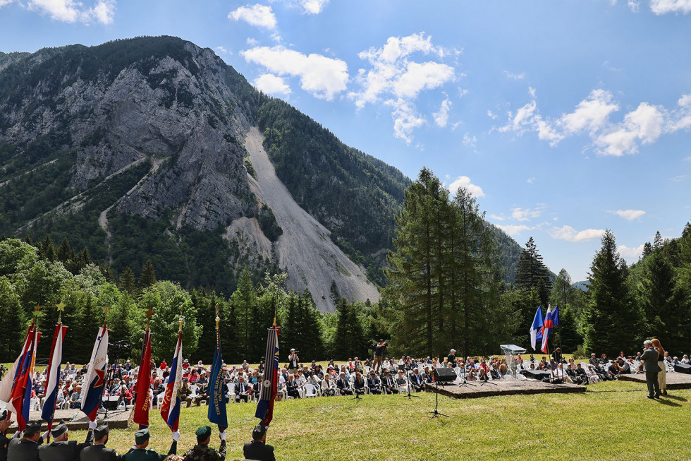 Prireditveni prostor z udeleženci od daleč, v ozadju vidne zastave, udeleženci in hribi