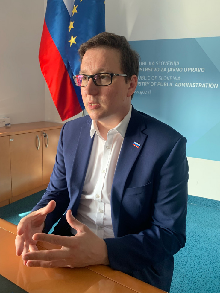 Boštjan Koritnik, sedeč za mizo, oblečen v suknjič, za njim vidna zastava slovenije in modo ozadje z logotipom ministrstva za javno upravo