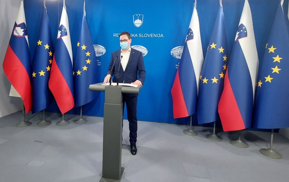 Minister stoji za govornico v tiskovnem centru vlade, zadaj modri pano Vlade Republike Slovenije, ob strani slovenska in EU zastava.