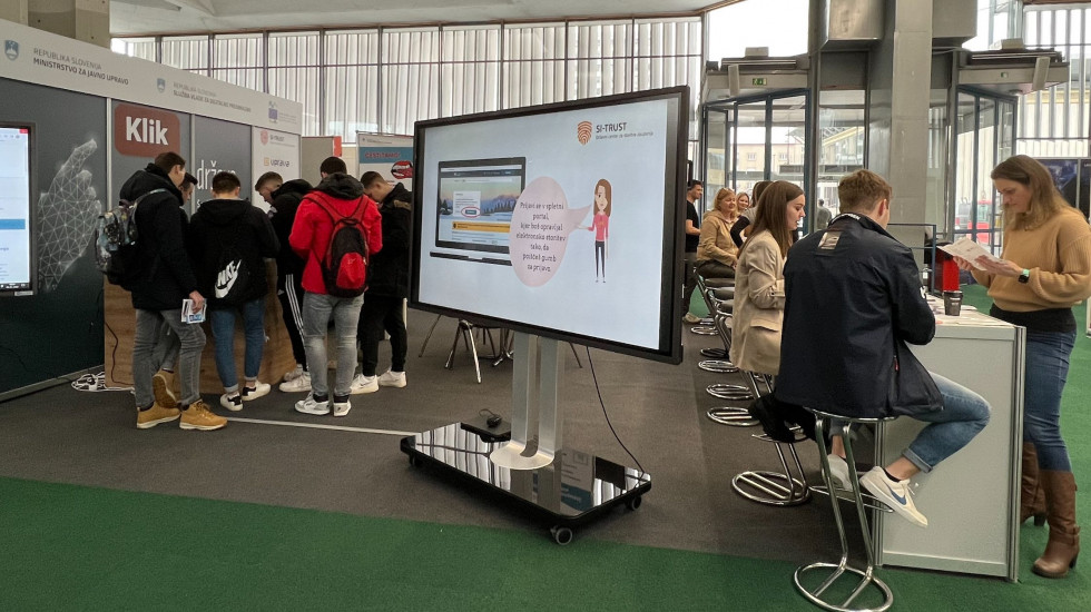Obiskovalci stojijo pred pultom in izpolnjujejo formularje, na razstavnem prostoru tudi veliki ekrani na katerem so video predstavitve, ki vabijo mlade, da se seznanijo z elektronskimi storitvami.