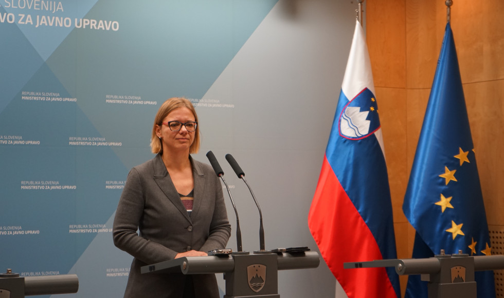 Ministrica stoječa za govornico, ob njej zastava Slovenije, za njo pano z logotipom ministrstva