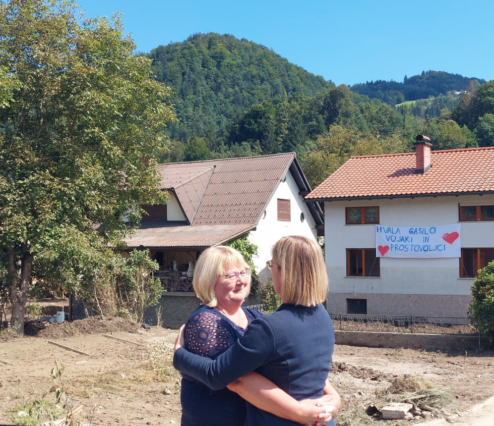 Ministrica in načelnica sta v objemu pred dvoriščem, kjer je razdejanje po poplavah