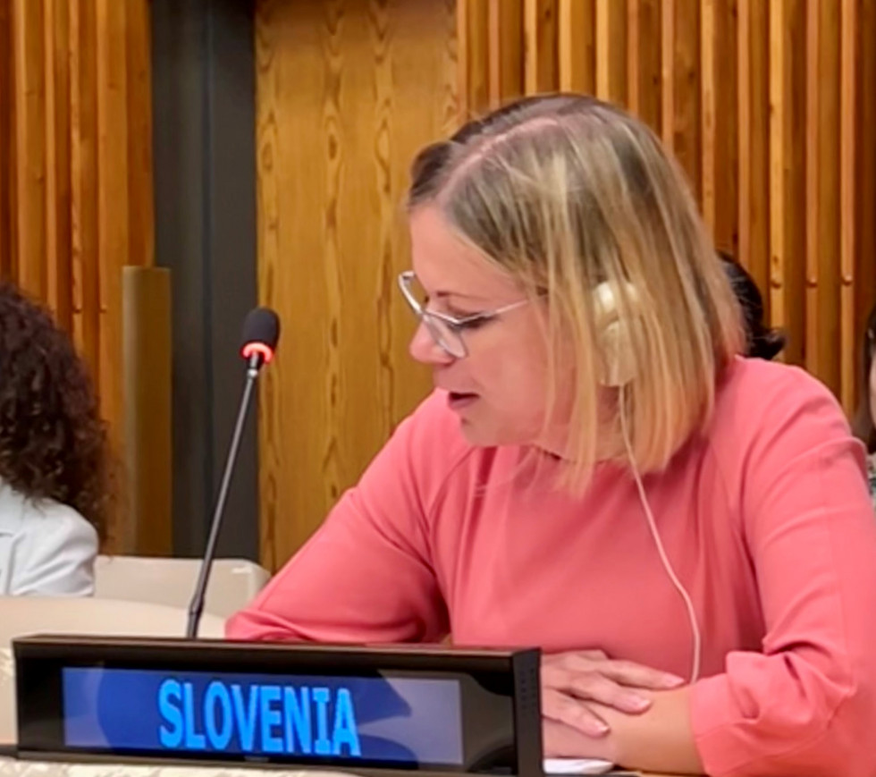 Ministrica sedi za mizo in govori v mikrofon, pred njo je majhen podolgovati ekranček z napisom Slovenia