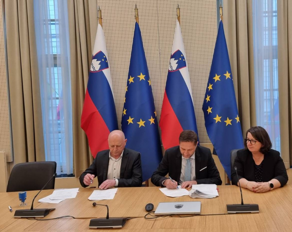 Ministra podpisujeta dogovor in anekse za mizo. Zadaj so zastave, slovenska in zastava EU