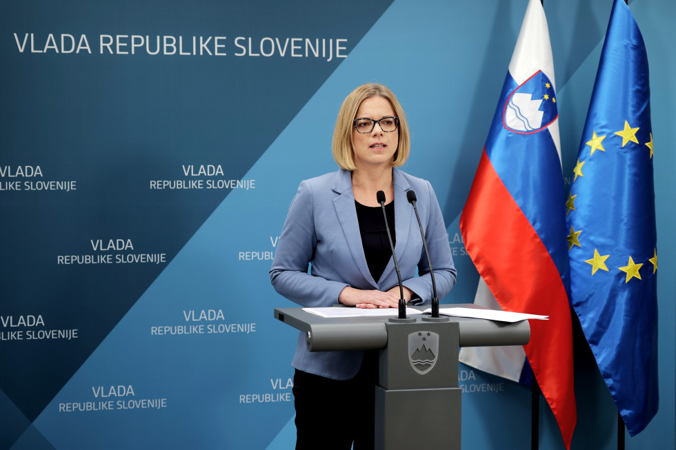 Ministrica stoječa za govornico, za njo ozadje Vlade RS, desno stoji slovenska zastava