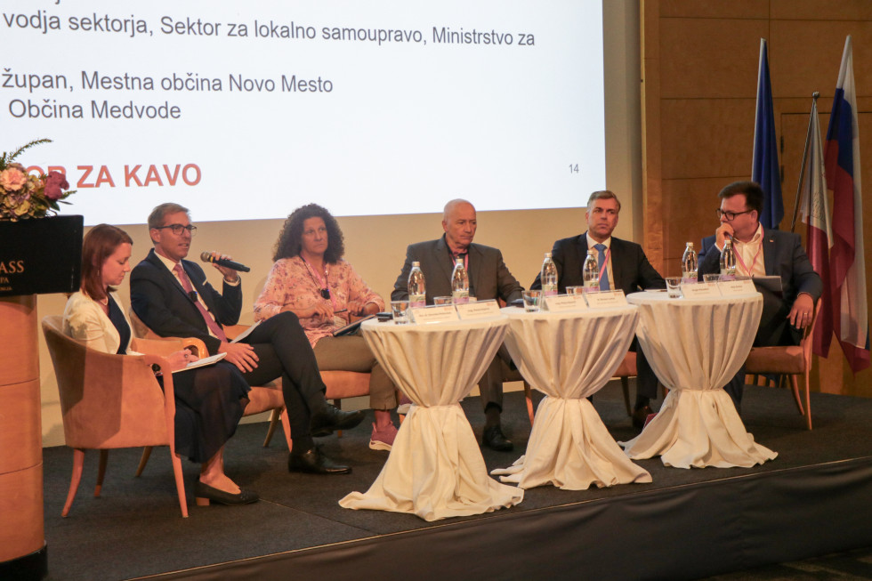 Dr. Roman Lavtar sodeluje v okrogli mizi z ostalimi sodelujočimi. Sedijo za mizo na odru. Zadaj so platno in zastave. in
