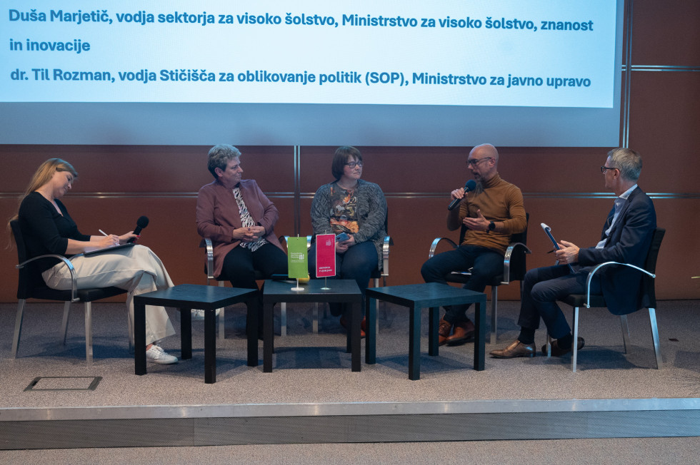 Na odru sedijo moderatorka in štirje govorci na okrogli mizi, mikrofon ima v roki dr. Til Rozman