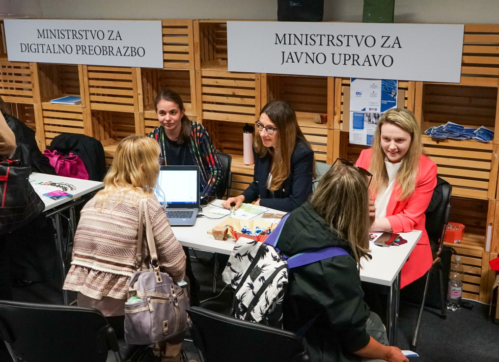 Sodelavke Ministrstva za javno upravo sedijo za mizo, kjer je razporejen promocijski material za udeležence sejma. Nasproti sedijo iskalci zaposlitve, ki se zanimajo glede zaposlitve v državni upravi.