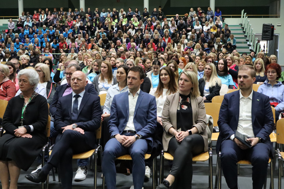 Ljudje sedijo v športni dvorani. V ospredju je prva vrsta, v kateri sedijo dve ženski in trije moški. 