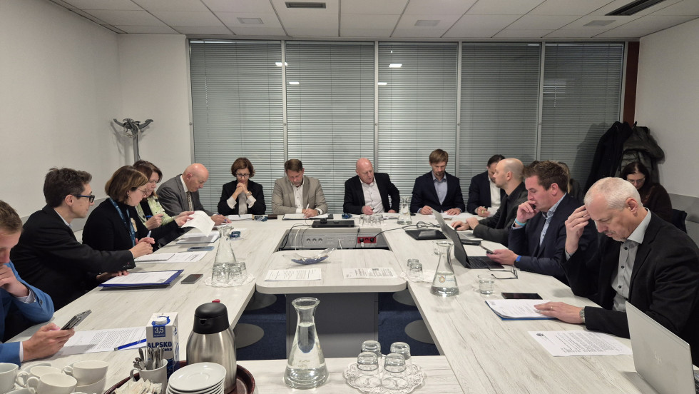 Okroglo omizje skupine posameznikov, ki gleda v gradivo, ki obravnavano v delovni skupini. Miza je bele barve, štirje glavni govorci sedijo s hrbti obrnjeni v okna.