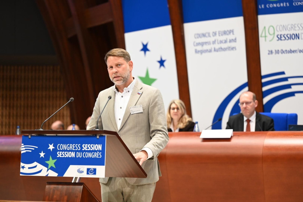 Državni sekretar stoji za govorniškim pultom in nagovarja udeležence v dvorani.