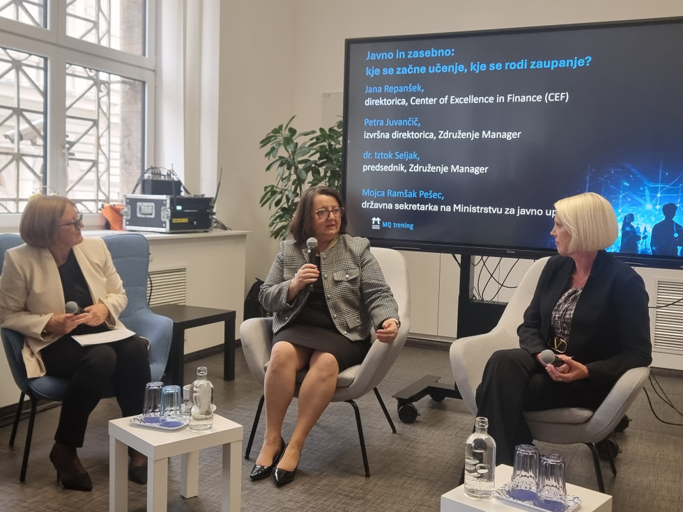 Tri sogovornice sedijo v pogovornem krogu pred zaslonom z naslovom razprave; ena izmed njih govori v mikrofon, preostali dve prisluhneta.